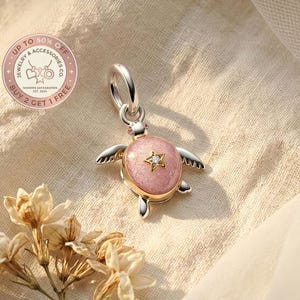 Può includere: Un ciondolo a forma di tartaruga argentata con guscio rosa e una stella dorata con una gemma trasparente. Il ciondolo ha piccole ali e fa parte di una collezione di gioielli. Su sfondo beige chiaro.