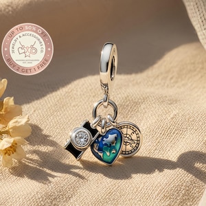 May include: A silver charm with three pendants: a camera with a clear stone, a blue and green heart with a map, and a compass. The charm is suspended from a silver loop. The image includes text that says "UP TO 50% OFF" and "BUY 2 GET 1 FREE".
