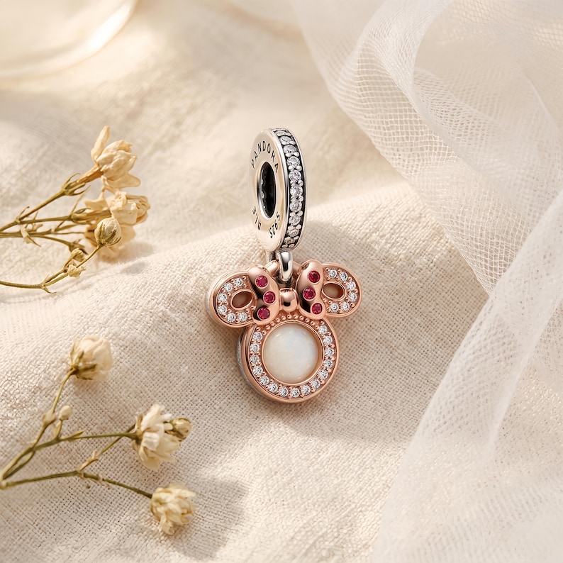 Puede incluir: Un dije colgante en tono oro rosa con forma de cabeza de rat&oacute;n, con una piedra circular blanca central rodeada de cristales transparentes. El lazo tiene piedras rojas y el pasador est&aacute; grabado con Pandora S925 y cristales transparentes.