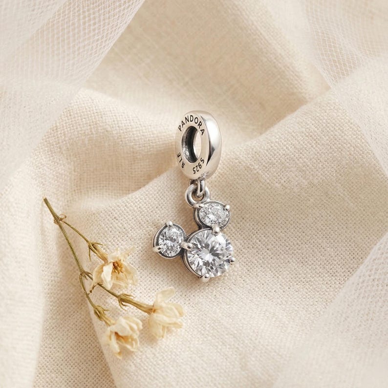 May include: A silver charm with a Mickey Mouse design. The charm features a large, round, clear gemstone for the face and smaller gemstones for the ears. The charm is suspended from a silver loop with the word "Pandora" engraved on it.