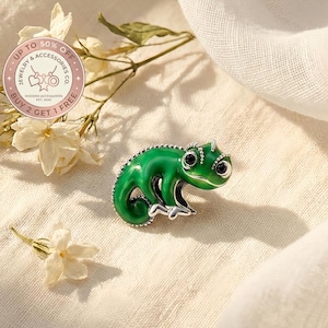 Puede incluir: Broche verde con forma de camaleón, con ojos negros y detalles plateados. La joya está sobre una superficie color crema con flores blancas y el texto "UP TO 50% OFF JEWELRY & ACCESSORIES CO." y "BUY 2 GET 1 FREE".