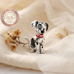 Puede incluir: Un charm de plata con forma de perro dálmata con manchas negras, un collar rojo y orejas negras. El charm está sobre un fondo suave y neutro con flores secas. El charm forma parte de una colección de joyas.