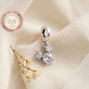 May include: A silver Pandora charm with a Mickey Mouse design, featuring three clear, round cubic zirconia stones. The charm is suspended from a silver loop. The image also includes a sprig of dried flowers and a circular logo with text.