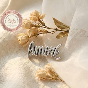 Peut inclure: Un pendentif argenté "Amore" en écriture cursive, posé sur un tissu blanc doux. Des fleurs séchées sont disposées autour du pendentif. L'image comprend également un logo circulaire avec le texte "UP TO 50% OFF JEWELRY & ACCESSORIES CO. BUY 2 GET 1 FREE".