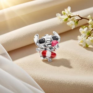 May include: A silver charm shaped like a character wearing black sunglasses and holding a pink ice cream cone. The character is wearing a red and white striped inner tube around its waist. The charm features detailed metallic textures and enamel.