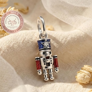 May include: A silver-colored charm in the shape of a nutcracker soldier. The charm has a blue hat with a star, red arms, and a detailed design. The charm is part of a jewelry collection.