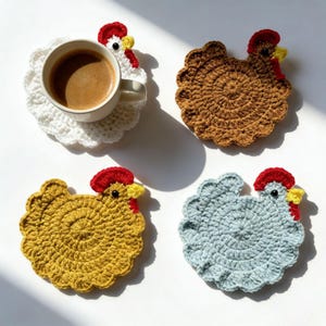 May include: Four round crochet coasters shaped like chickens in white, brown, mustard yellow, and light blue. Each coaster features a red comb, a yellow beak, and a black eye. A white ceramic mug filled with coffee sits on the white coaster.