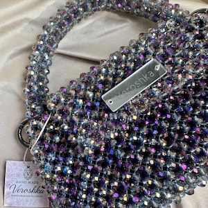 May include: A handbag constructed from iridescent, multi-faceted beads in shades of deep purple, blue, and silver. The bag features a rounded handle and a rectangular silver-toned tag engraved with the text Veroshka, plus a square paper tag.