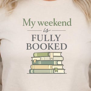 May include: A cream-coloured t-shirt featuring the text "My weekend is FULLY BOOKED" in green and black. The design includes a stack of books in green and beige with bookmarks. The t-shirt is a casual style.