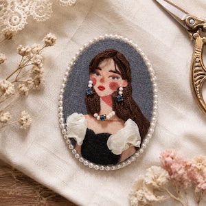 Puede incluir: Un broche de retrato bordado de forma ovalada que muestra una figura con cabello largo castaño, vistiendo un top negro con mangas blancas con volantes, un collar de perlas con una piedra azul central y pendientes a juego, enmarcado por perlas blancas.