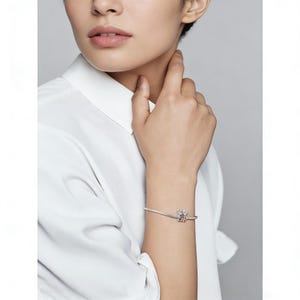 Puede incluir: Una pulsera de plata con un charm que presenta un peque&ntilde;o dise&ntilde;o detallado con detalles en rojo y blanco. La pulsera se lleva en la mu&ntilde;eca, con una camisa blanca en el fondo.