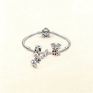 Puede incluir: Una pulsera de plata con varios abalorios. Los abalorios incluyen un carruaje de Cenicienta, una campana de Navidad, un abalorio con dos ratones de dibujos animados y un abalorio con un dise&ntilde;o rojo y blanco. La pulsera est&aacute; sobre un fondo blanco.