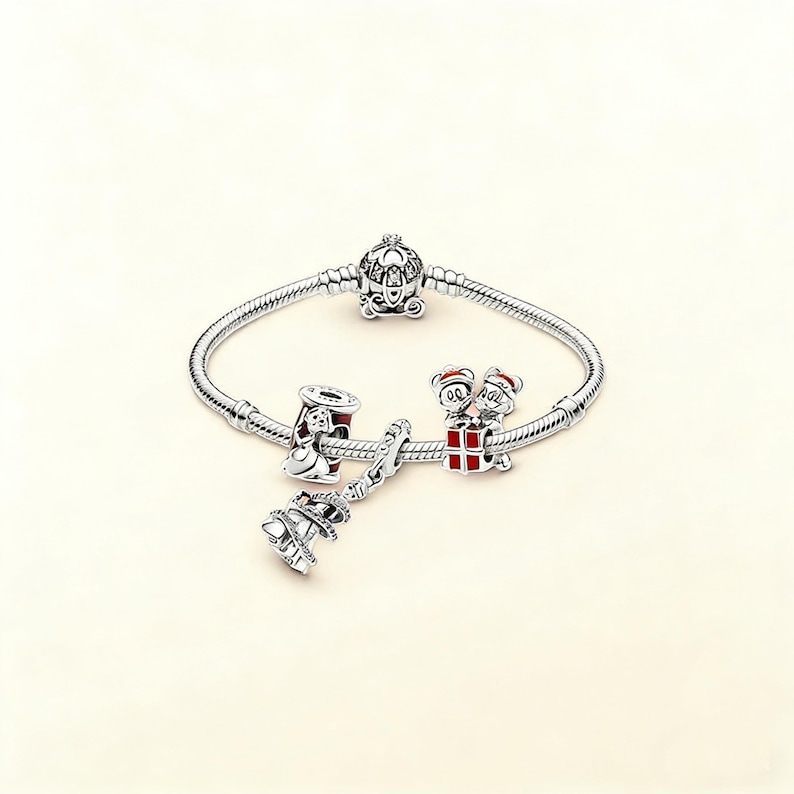 Puede incluir: Una pulsera de plata con varios abalorios. Los abalorios incluyen un carruaje de Cenicienta, una campana de Navidad, un abalorio con dos ratones de dibujos animados y un abalorio con un dise&ntilde;o rojo y blanco. La pulsera est&aacute; sobre un fondo blanco.