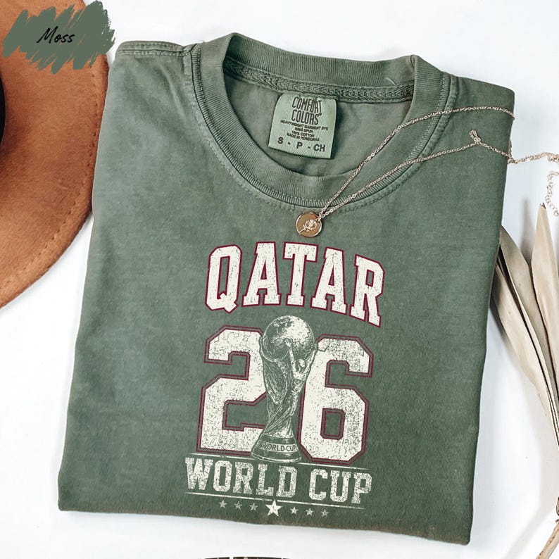 A green t-shirt with the text "QATAR 26 WORLD CUP" printed on it, along with a trophy icon.