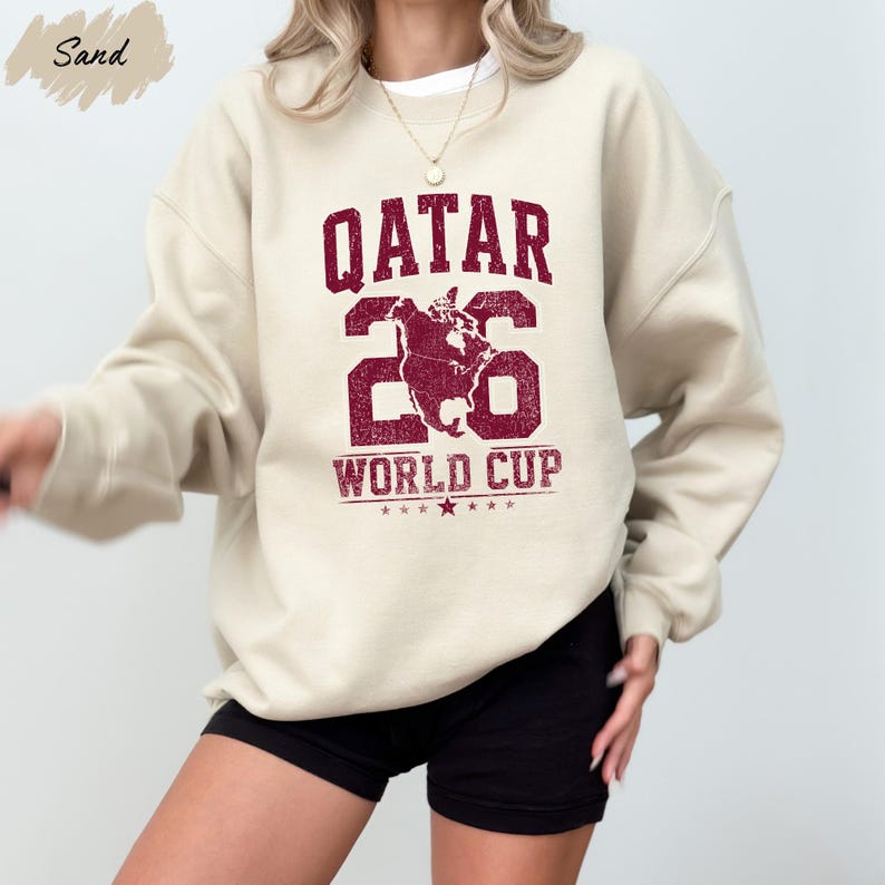 A person wearing a white sweatshirt with a maroon graphic and text that says "QATAR 26 WORLD CUP".
