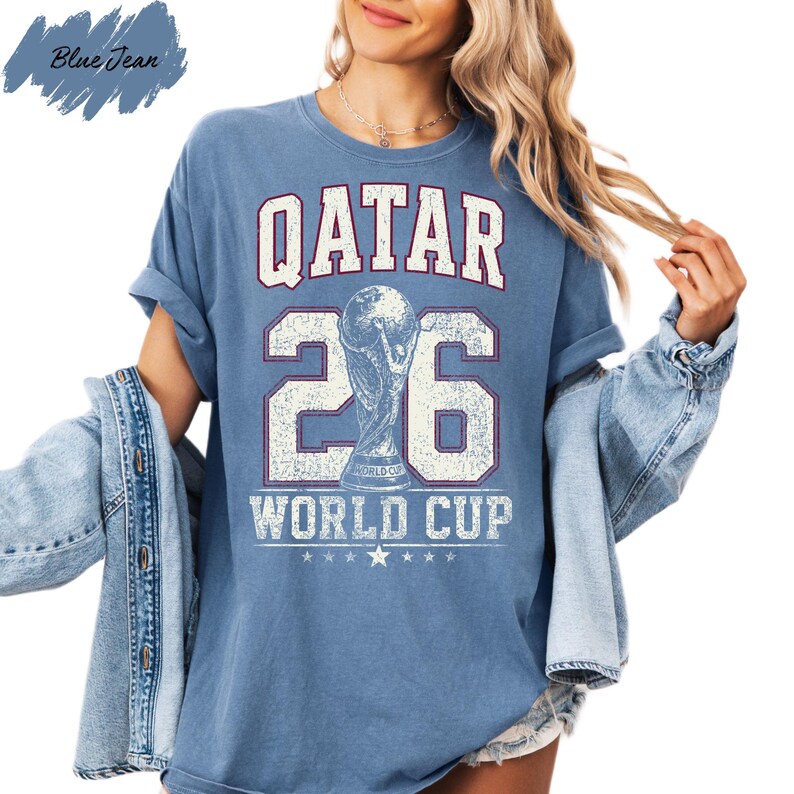 A woman wearing a blue t-shirt with the text "QATAR 26 WORLD CUP" printed on it, along with a trophy icon. She is also wearing a denim jacket and has her hair in a ponytail.