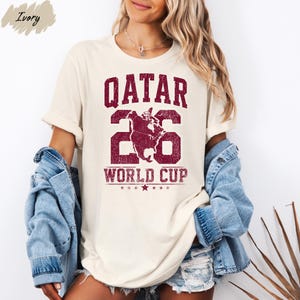 A woman wearing a white t-shirt with the text "QATAR 26 WORLD CUP" printed on it, along with a graphic of a football. She is also wearing denim shorts and a denim jacket.
