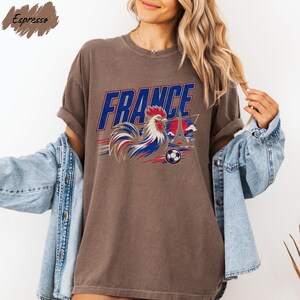 A woman wearing a brown t-shirt with a graphic of a rooster and the word "FRANCE" printed on it, along with a denim jacket.