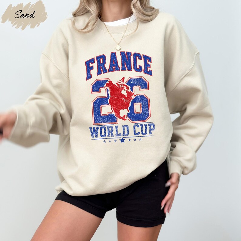 A person wearing a beige sweatshirt with a graphic design featuring the text "FRANCE 26 WORLD CUP" and a map of the world.