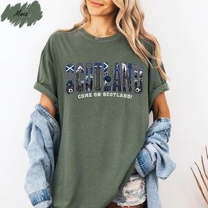 A woman wearing a green t-shirt with the word "SCOTLAND" printed on it, along with the text "COME ON SCOTLAND!" printed below. She is also wearing a denim jacket and denim shorts.