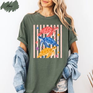 A woman wearing a green t-shirt with a colorful graphic of three sea turtles and stars on it, along with a denim jacket.