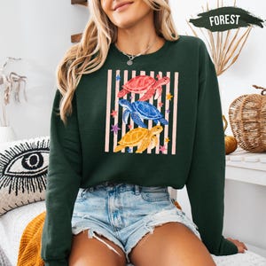 A woman wearing a green sweatshirt with a graphic of colorful sea turtles and a striped pattern, paired with denim shorts.