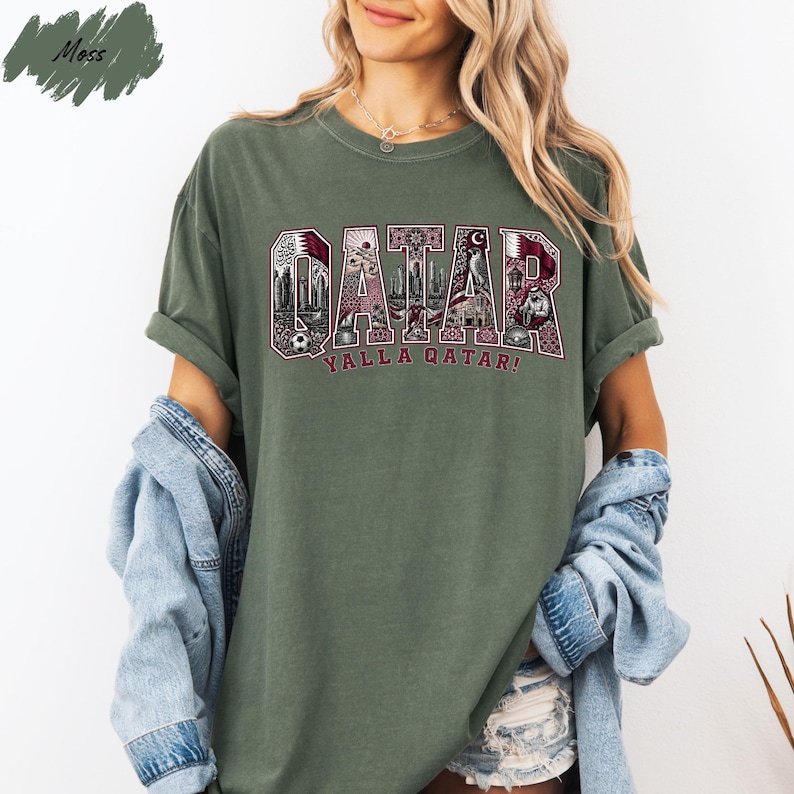 A woman wearing a green t-shirt with the word "QATAR" printed on it, along with the phrase "YALLA QATAH!" printed below. She is also wearing a denim jacket and denim shorts.