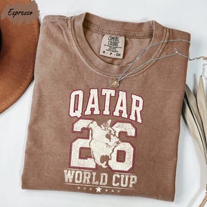 A brown t-shirt with the text "QATAR 26 WORLD CUP" printed on it, along with a map of the world. The t-shirt is folded and placed on a white surface, with a necklace and a hat visible in the background.