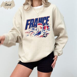 A person wearing a beige sweatshirt with a graphic design featuring a rooster and the word "FRANCE" printed on it.