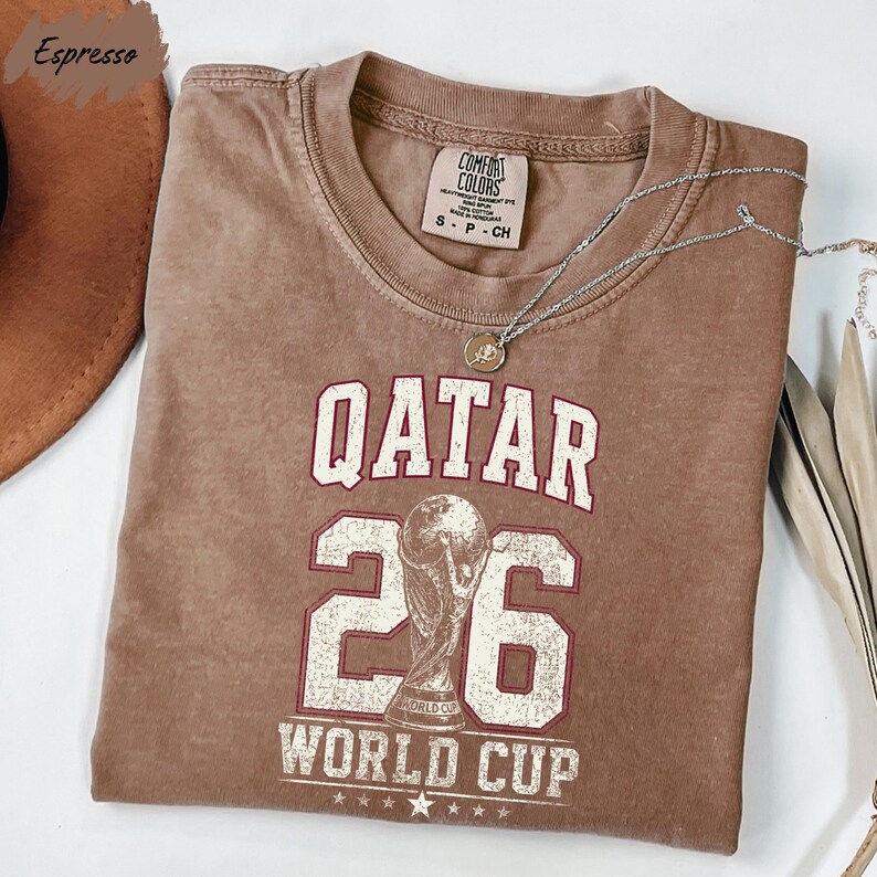 A brown t-shirt with the text "QATAR 26 WORLD CUP" printed on it, along with a trophy icon.