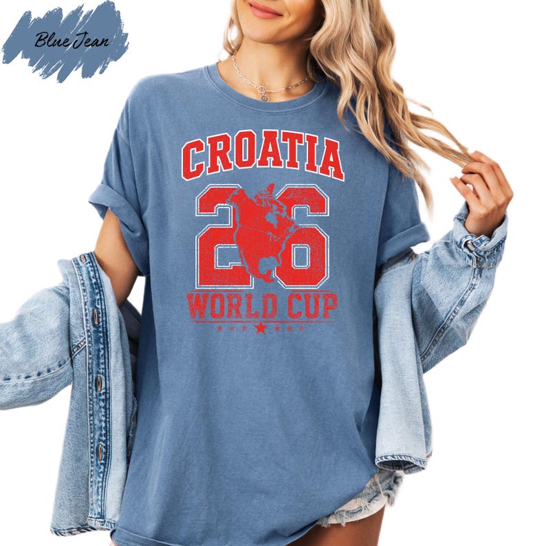 A woman wearing a blue t-shirt with the text "Croatia 26 World Cup" printed on it, along with a graphic of a dog. She is also wearing a denim jacket and has her hair styled in loose waves.