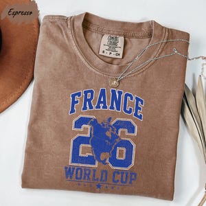 A brown t-shirt with the text "FRANCE 26 WORLD CUP" printed on it, along with a number "26" and a graphic of a globe.