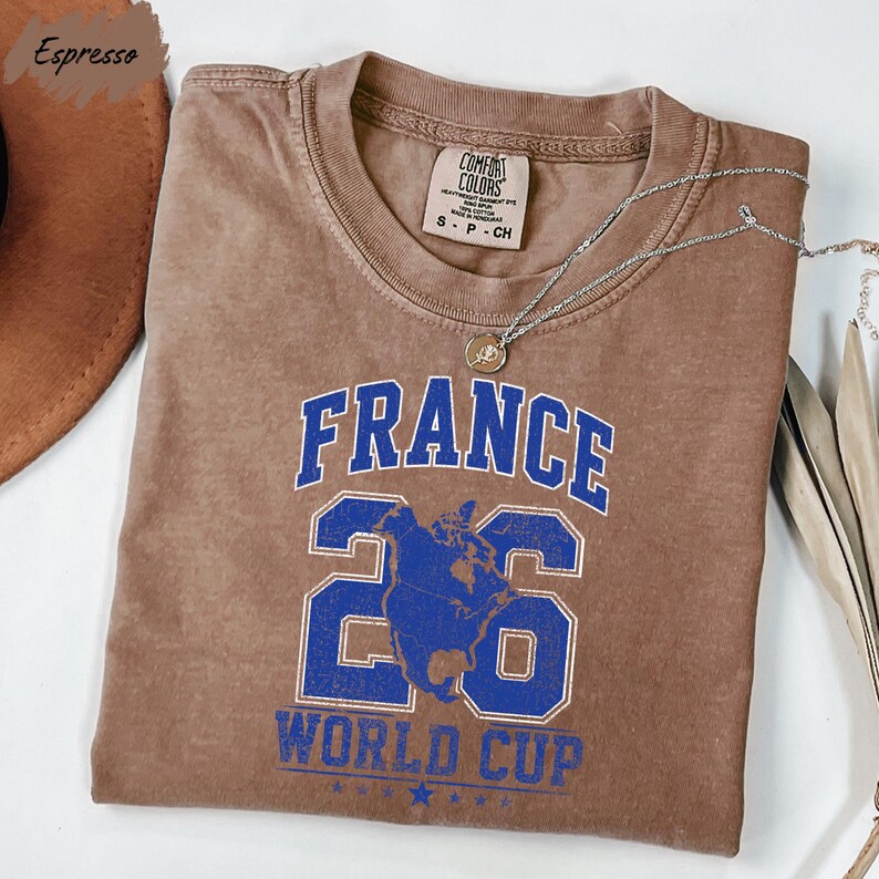 A brown t-shirt with the text "FRANCE 26 WORLD CUP" printed on it, along with a number "26" and a graphic of a globe.