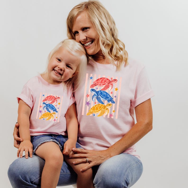 A smiling woman wearing a pink t-shirt with a design of colorful sea creatures and a child wearing a matching pink t-shirt.