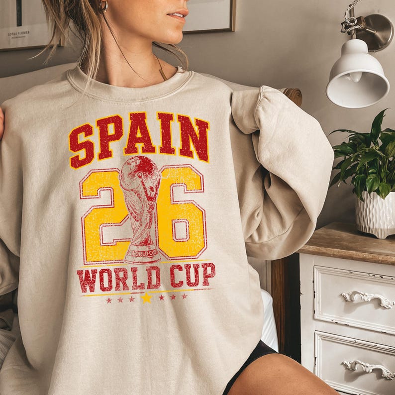 A person wearing a beige sweater with a graphic design that says "SPAIN 26 WORLD CUP" and features a trophy.