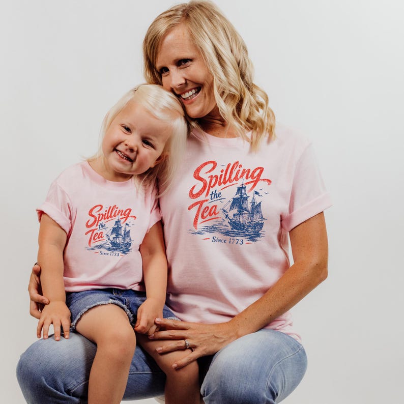 A smiling woman wearing a pink t-shirt with a graphic of a ship and the text "Spilling the Tea Since 1773" is holding a young girl in a matching pink t-shirt.