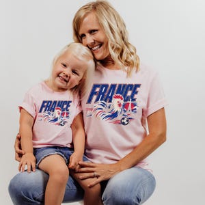 A smiling woman wearing a pink t-shirt with the word "FRANCE" and a graphic of a rooster, holding a young girl in her lap.