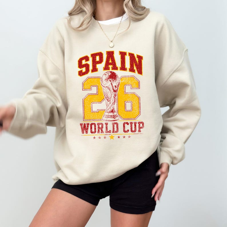 A person wearing a white sweatshirt with a graphic design that says "SPAIN 26 WORLD CUP" and a trophy.