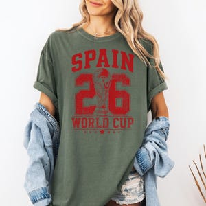 A woman wearing a green t-shirt with the text "SPAIN 26 WORLD CUP" printed on it, standing against a white background.