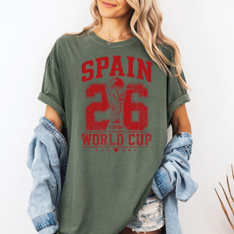 A woman wearing a green t-shirt with the text "SPAIN 26 WORLD CUP" printed on it, standing against a white background.