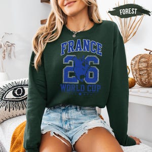 A woman wearing a green sweatshirt with the text "FRANCE" and "WORLD CUP" printed on it, along with the number "26" and a graphic of a hand. She is also wearing denim shorts and a necklace.