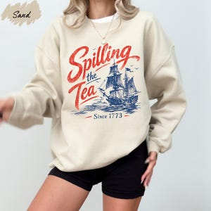 A person wearing a beige sweatshirt with a graphic design featuring a ship and the text "Spilling the Tea Since 1773".