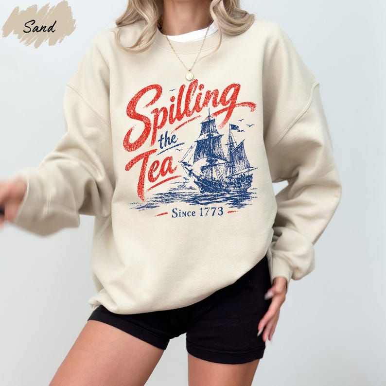 A person wearing a beige sweatshirt with a graphic design featuring a ship and the text "Spilling the Tea Since 1773".