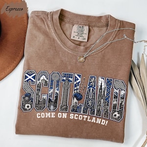 a brown t-shirt with the word "SCOTLAND" written on it in a stylized font, along with the phrase "COME ON SCOTLAND!" printed below. The t-shirt is laid out on a white surface, and there is a necklace visible in the background.