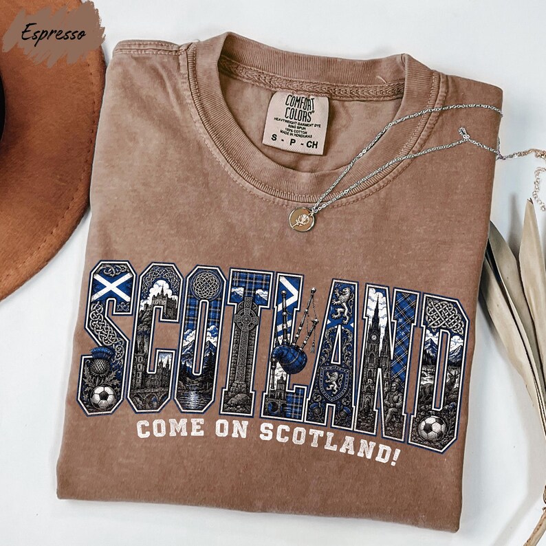 a brown t-shirt with the word "SCOTLAND" written on it in a stylized font, along with the phrase "COME ON SCOTLAND!" printed below. The t-shirt is laid out on a white surface, and there is a necklace visible in the background.