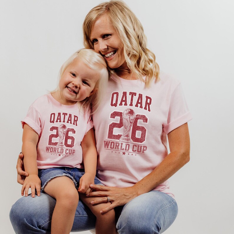 A smiling woman wearing a pink t-shirt with the number "26" and the text "QATAR 26 WORLD CUP" is holding a young girl in a matching pink t-shirt.