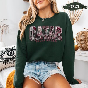 A woman wearing a green sweatshirt with the word "Qatar" printed on it, paired with denim shorts, sits in a room with various decorative items in the background.