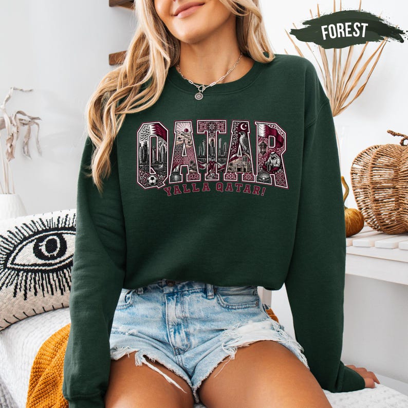 A woman wearing a green sweatshirt with the word "Qatar" printed on it, paired with denim shorts, sits in a room with various decorative items in the background.