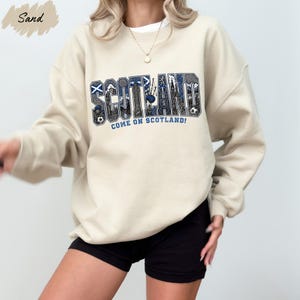 A person wearing a beige sweatshirt with the word "SCOTLAND" printed on it, along with the text "COME ON SCOTLAND!" printed below.