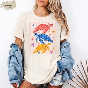A woman wearing a white t-shirt with a colorful graphic of three sea turtles and stars, paired with denim shorts and a denim jacket.